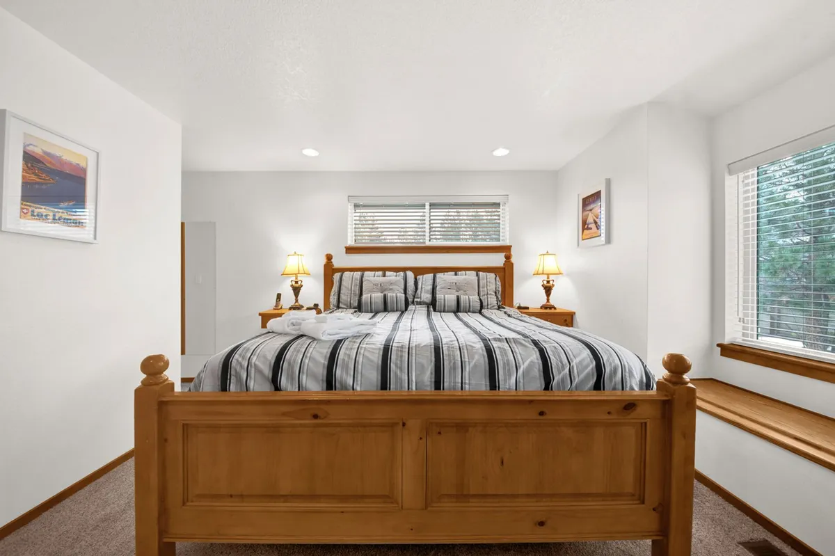 A serene bedroom featuring a large wooden bed with striped bedding, nightstands with lamps, a window letting in natural light, and framed artwork. The space feels comfortable and restful, ideal for unwinding after a busy day.