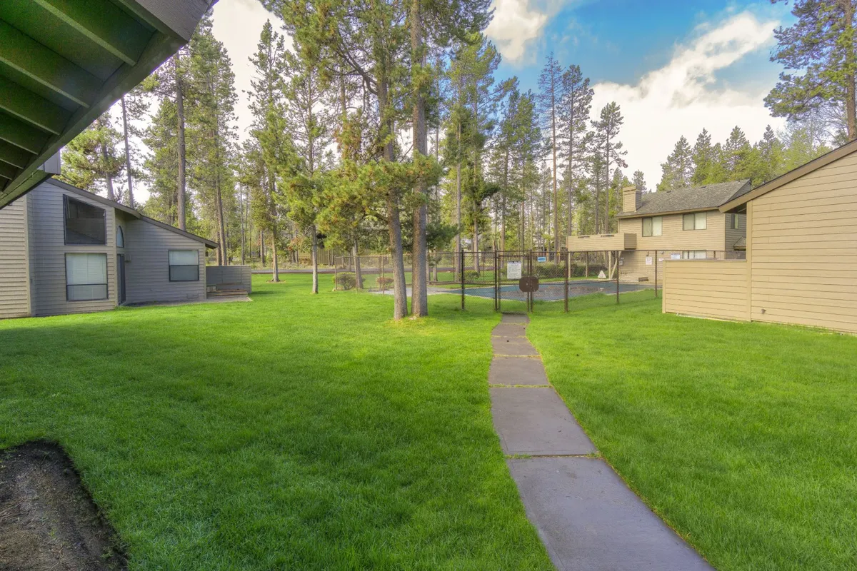 A lush, green lawn surrounded by tall trees and inviting pathways, with buildings in the distance. People can be seen walking, enjoying picnics, and playing outdoor games, creating a vibrant community atmosphere.