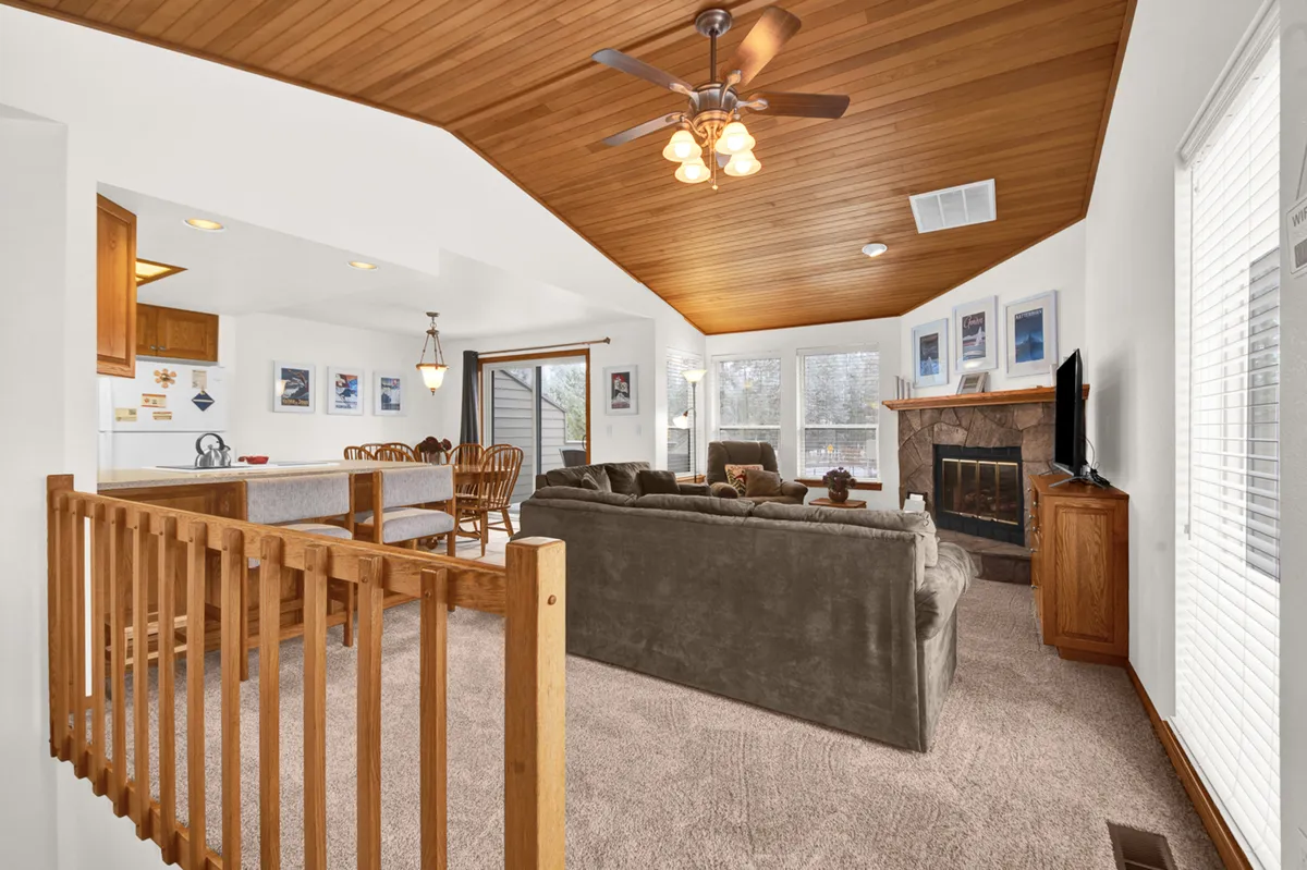 A spacious living and dining area is bathed in natural light from large windows. The room features a cozy fireplace, plush sofas, a large flat-screen TV, and an adjacent dining area with a wooden table, all under a warm wooden ceiling. The overall atmosphere is open and inviting, ideal for gatherings and relaxation.