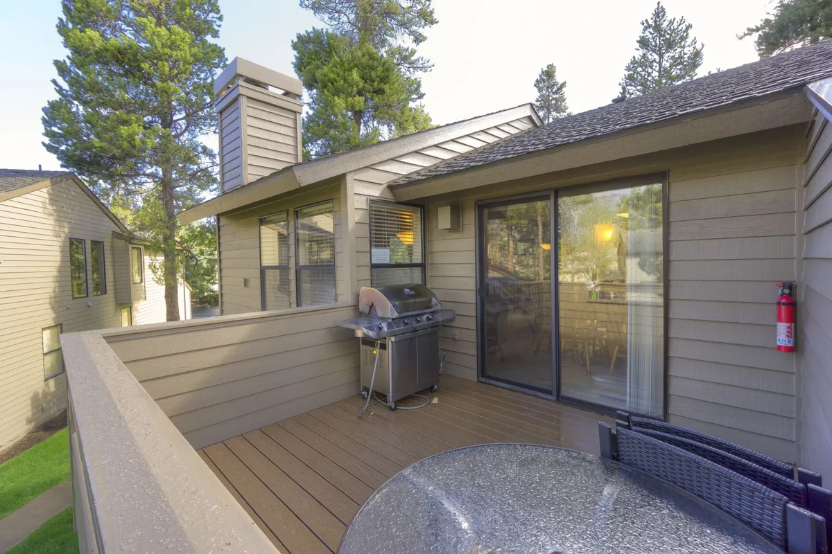 An exterior view of a spacious deck furnished with a table and surrounded by trees. A grill sits nearby, and a sliding glass door connects the deck to the interior, suggesting easy movement between indoor and outdoor living spaces. The deck invites al fresco dining and relaxation under the sky.