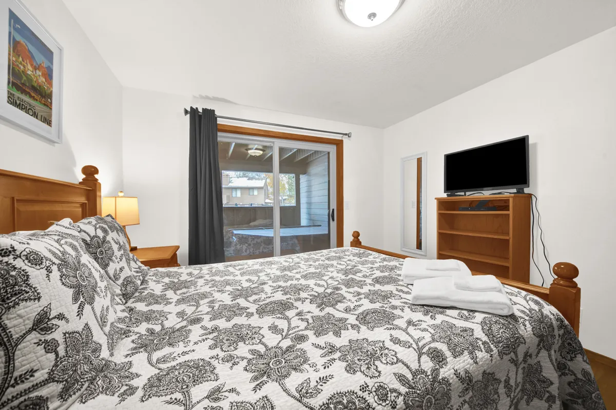 A cozy bedroom with a bed adorned with patterned bedding, a sliding glass door offering outdoor access, and a flat-screen TV on the dresser. The room receives gentle morning light and feels warm and inviting.